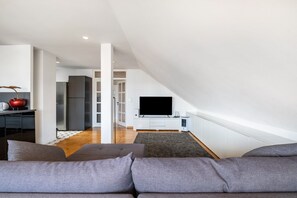 Una sala de estar moderna con techo alto, un televisor de pantalla plana montado en un mueble blanco, un sofá gris y un comedor con cocina al fondo.