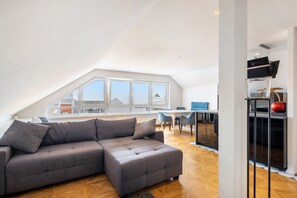 A modern loft apartment with a large gray sectional sofa, a dining area with a white table and blue chairs, and a kitchen area with a black cabinet and a red apple on top.