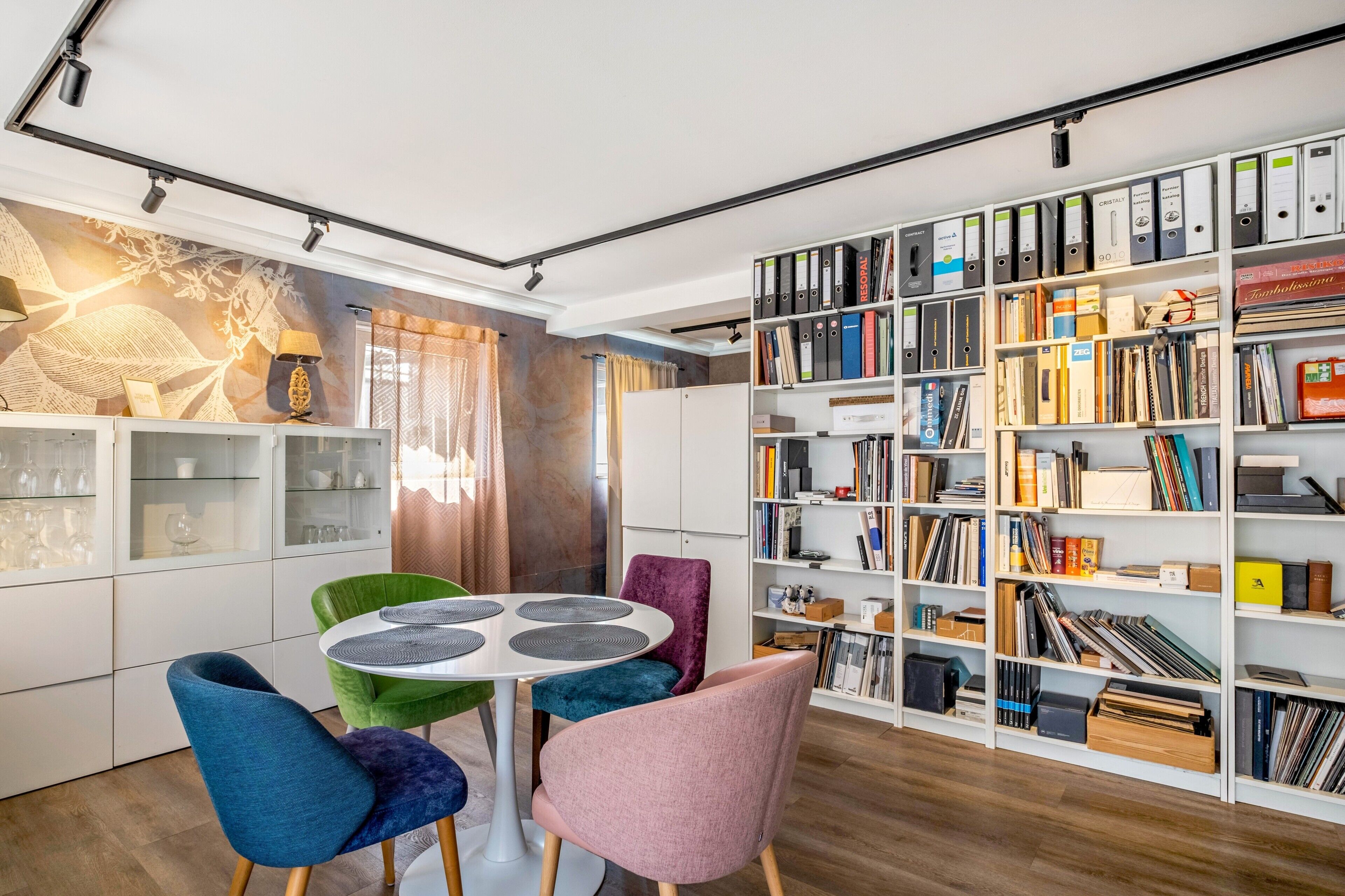 A modern interior with a round table surrounded by colorful chairs, a bookshelf filled with books and binders, and a decorative wall with a geometric pattern.