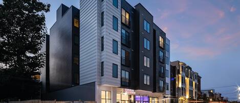 A multi-story hotel with a modern architectural design, featuring a prominent corner building and a ground-level shop.