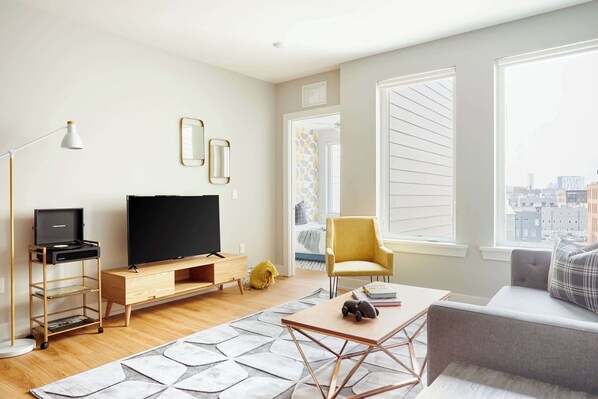 Un salon moderne doté d’un téléviseur à écran plat, d’un meuble de rangement en bois, d’un fauteuil jaune, d’un canapé gris et offrant une vue sur la ville grâce à de grandes fenêtres.