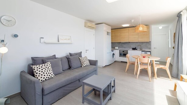 Un appartement moderne avec un canapé gris, une petite table, une cuisine dotée de meubles en bois et un coin repas avec des chaises.
