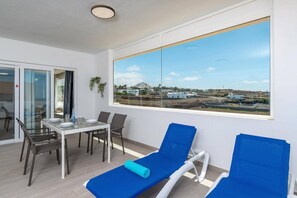 Ein Zimmer mit Esstisch, Stühlen, einem blauen Lounge-Sessel und einem großen Fenster mit Blick auf Gebäude und eine Landschaft.