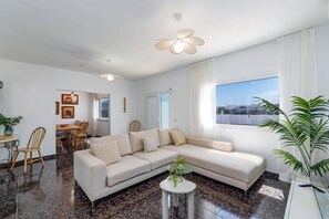 A modern living room with a sectional sofa, a dining area with wooden chairs, a ceiling fan, and a large window with a view. - Villa Camino del Volcan by Villa Plus (Mácher)