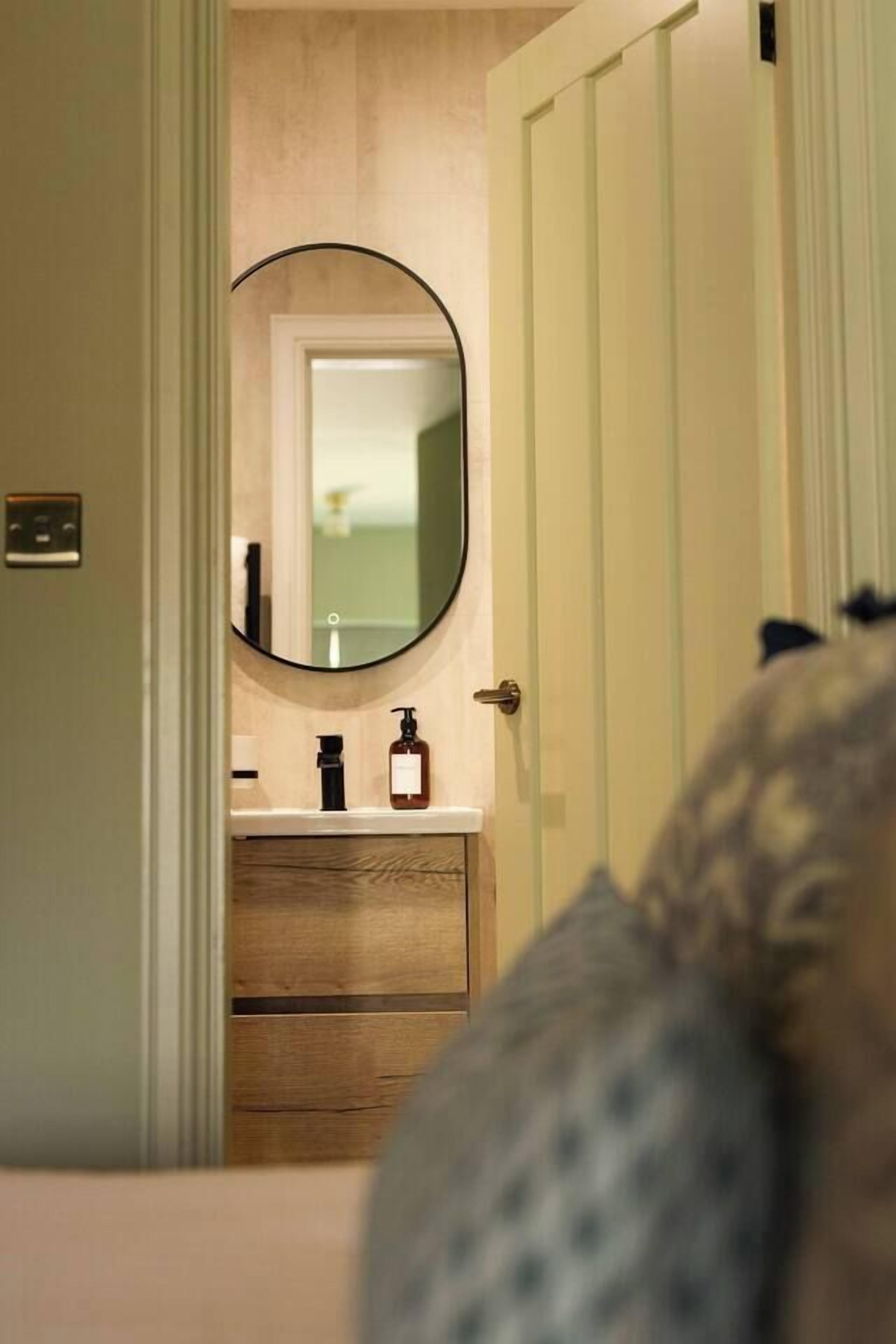 A bathroom with a wooden vanity, a round mirror, and a door leading to another room.