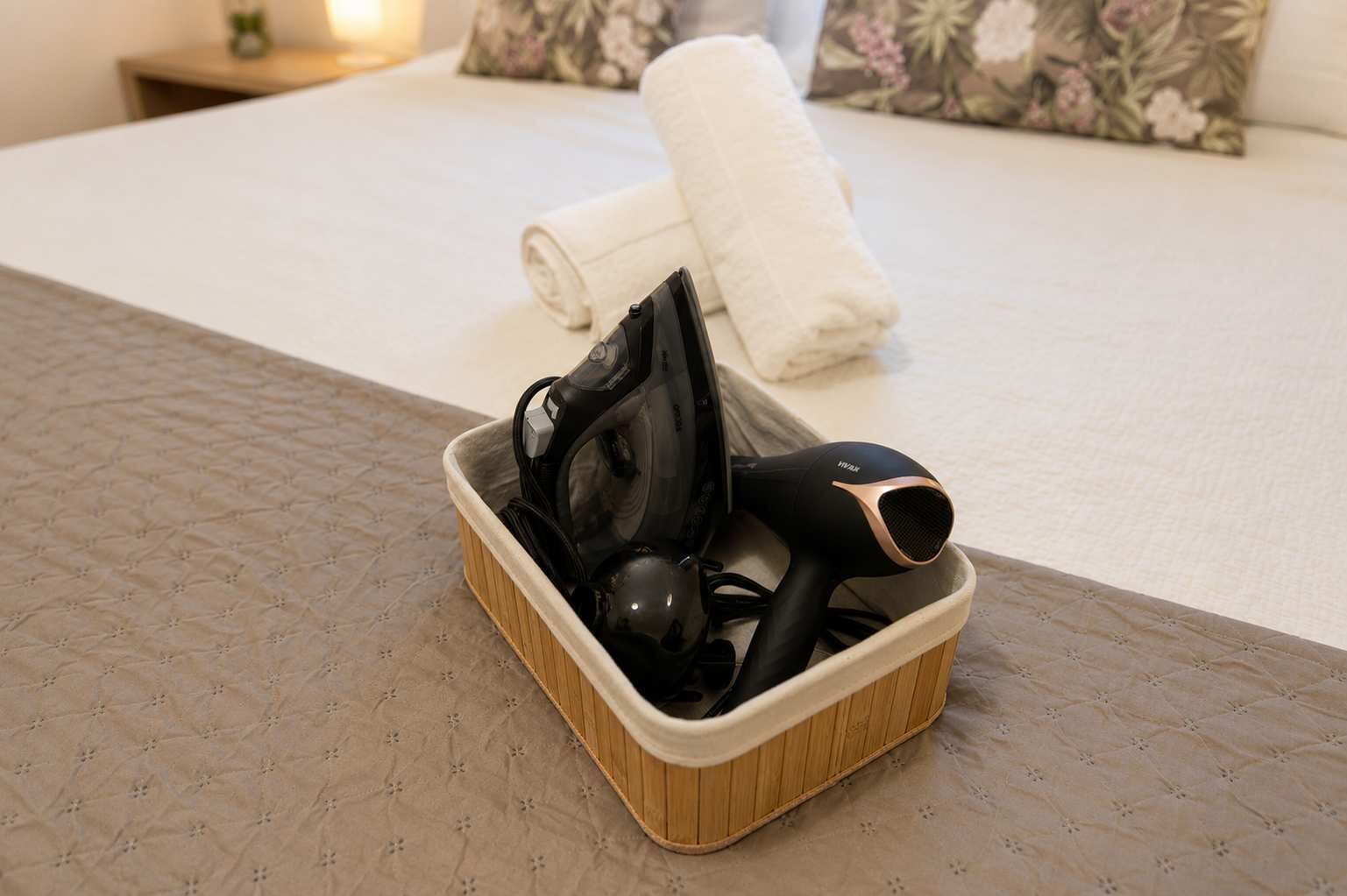Ein Hotelzimmer mit einem ordentlich bezogenem Bett, einem kleinen Holfnachtkästchen mit einer Lampe sowie einem Korb mit einem Föhn und weiteren Gegenständen.
