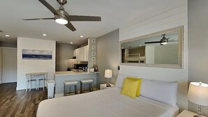 A hotel room with a bed, a ceiling fan, a nightstand, a lamp, a mirror, a kitchenette, and a seating area with stools. - 🌴Sandpiper Cove 8242~Steps to the beach- 5 Pools + 3 Hot Tubs~ Golf Course ⛳ (Destin)