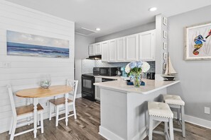 A modern kitchen with white cabinetry, a wooden dining table, and a small island with bar stools. - 🌴Sandpiper Cove 8242~Steps to the beach- 5 Pools + 3 Hot Tubs~ Golf Course ⛳ (Destin)