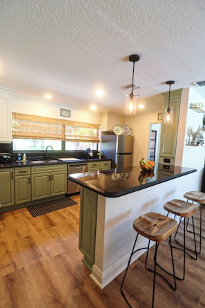 A kitchen with wooden flooring, a central island with stools, stainless steel appliances, and a window with blinds. - Heated pool, Putting Green, propane fire pit, outdoor shower waterfront home 4 U (Palm Beach Gardens)