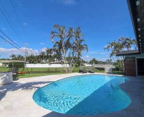 A swimming pool with a curved edge, surrounded by a concrete deck. There are lounge chairs and a swing set in the background. - Heated pool, Putting Green, propane fire pit, outdoor shower waterfront home 4 U (Palm Beach Gardens)