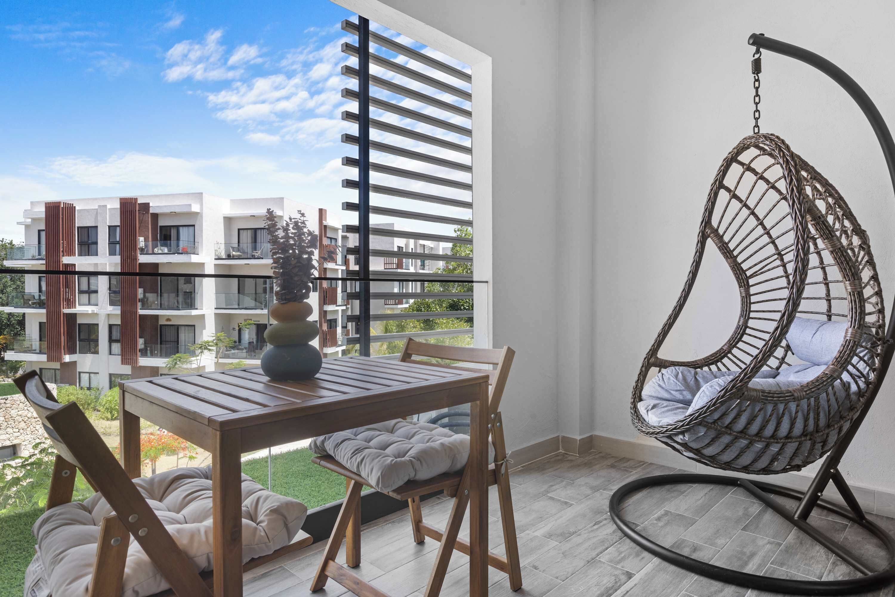 Un balcon avec une table en bois, des chaises, un fauteuil suspendu et une vue sur des immeubles modernes.