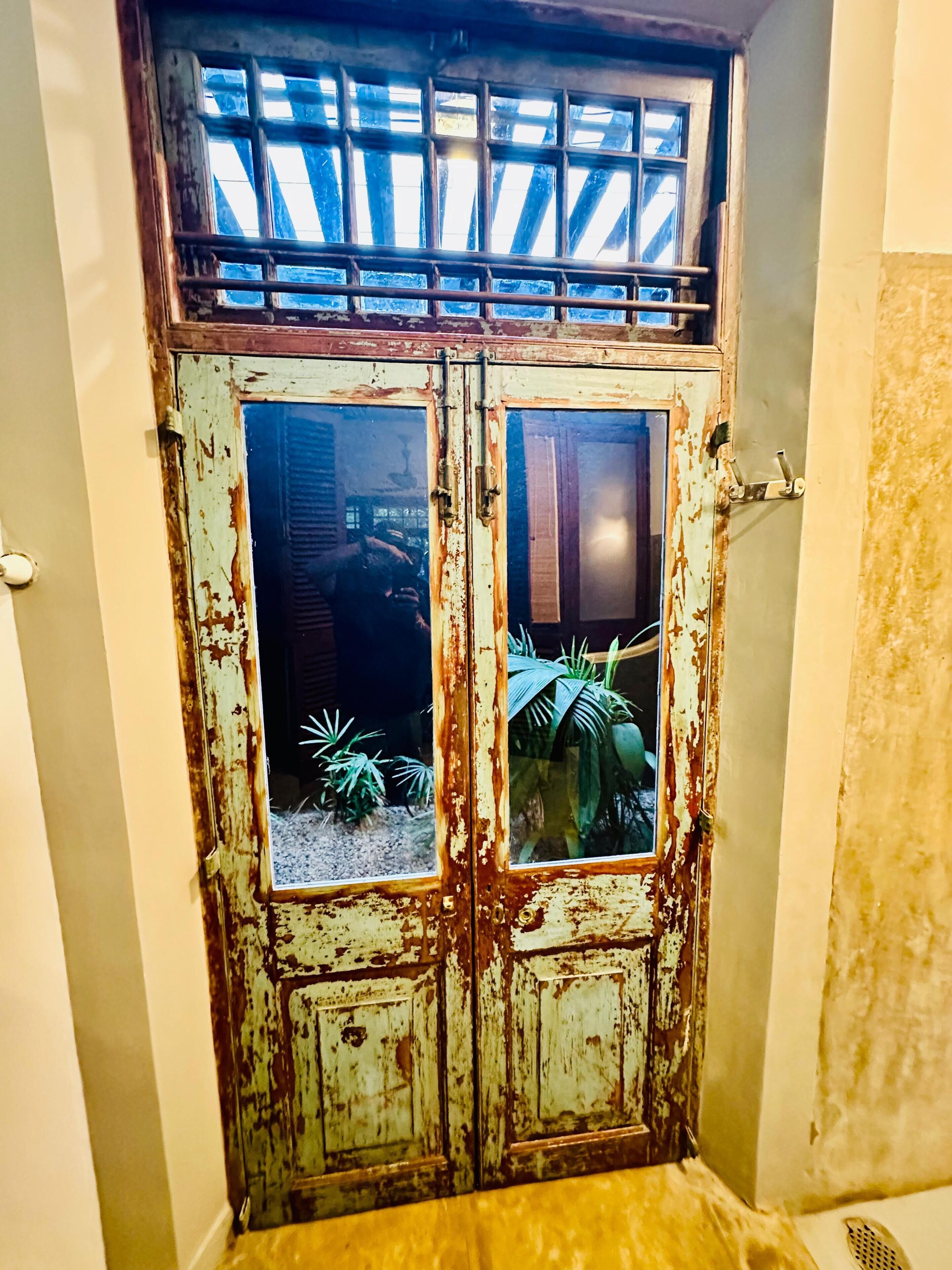 A weathered double door with a window, a metal grid above it, and a reflection of a person.