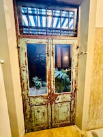 Puerta doble de madera gastada con un ventanal y una rejilla metálica encima, con el reflejo de una persona.