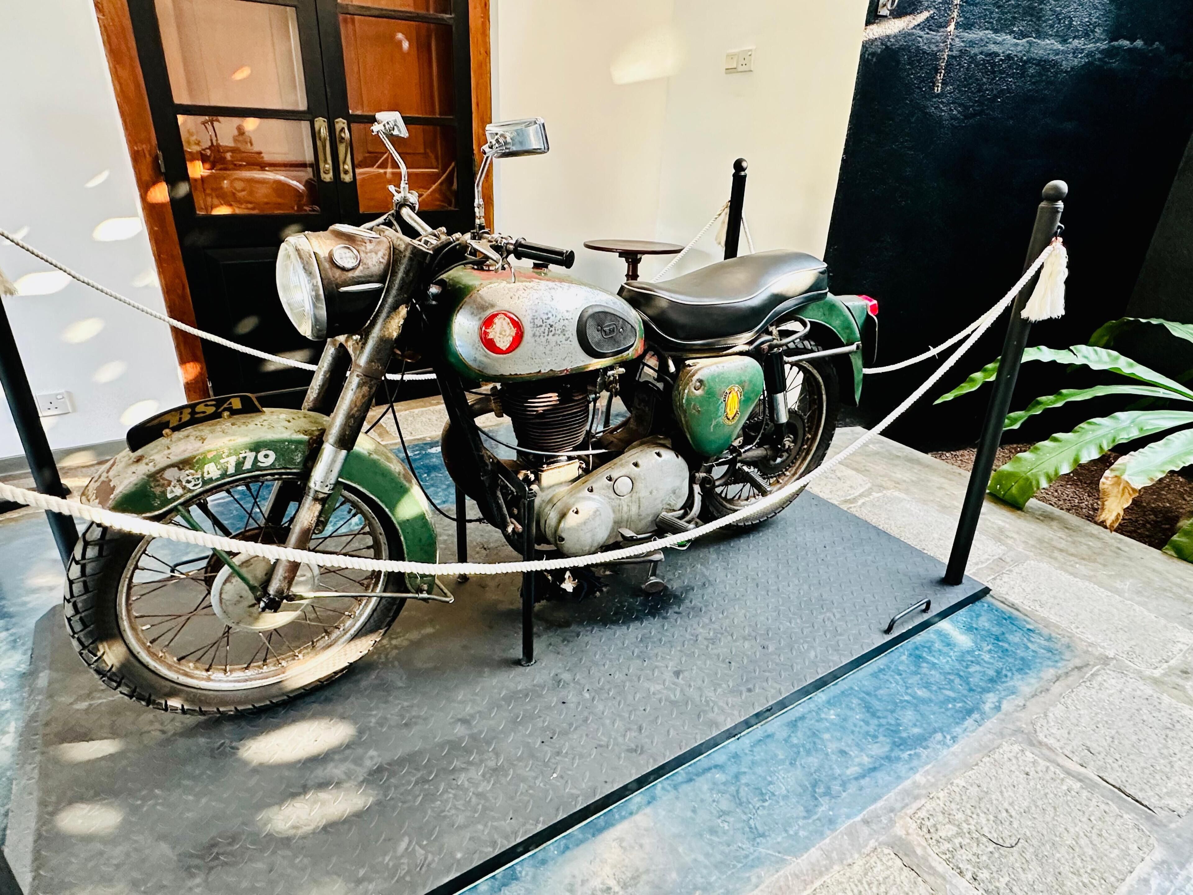 Una motocicleta antigua expuesta, asegurada con cuerdas, en una habitación con piso estampado y una puerta al fondo.