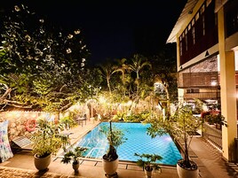 Vista nocturna de una piscina rodeada de plantas en macetas y árboles, con un edificio a un lado.