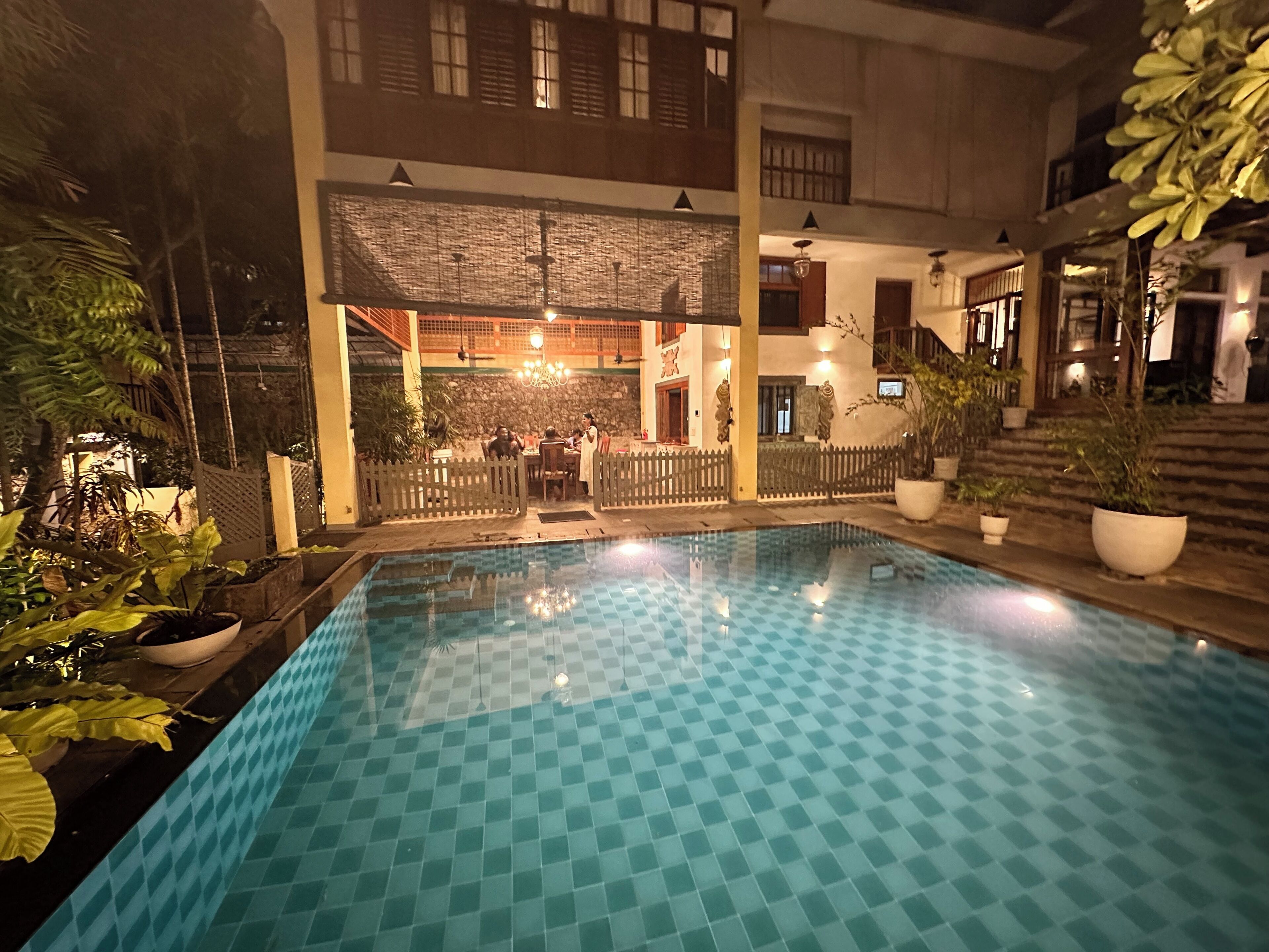 Una piscina con diseño ajedrezado, rodeada de plantas en macetas y un edificio con varios pisos y balcones.