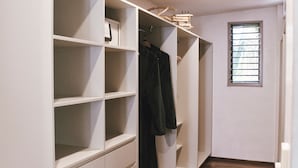 Ein begehbarer Kleiderschrank mit mehreren Regalen und Kleiderstangen, ein Fenster mit Jalousien und ein Holzboden.