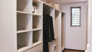 Ein begehbarer Kleiderschrank mit mehreren Regalen und Kleiderstangen, ein Fenster mit Jalousien und ein Holzboden.