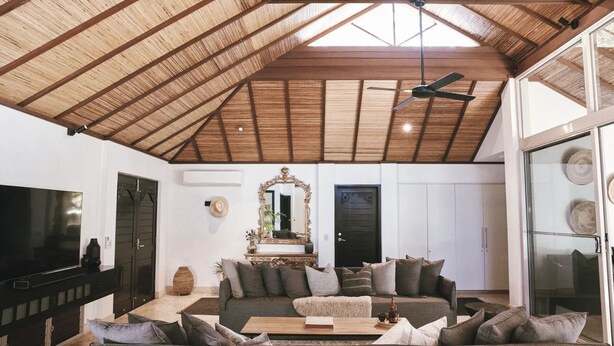 Ein Wohnzimmer mit hoher Decke, einem Deckenventilator, einem großen Flachbildfernseher, einer Couch mit Kissen, einem Couchtisch und einem dekorativen Spiegel.