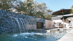 Ein Schwimmbecken mit Wasserfall, eine Lounge-Bereich mit Gartenmöbeln und eine Kachelwand.