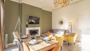 A dining area with a wooden table set for four, a hanging chair, and a sofa with cushions. A flat-screen TV is mounted above a fireplace. - Boutique Eastbourne Gem (Eastbourne)