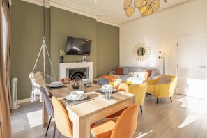 A dining area with a wooden table set for four, a hanging chair, and a sofa with cushions. A flat-screen TV is mounted above a fireplace. - Boutique Eastbourne Gem (Eastbourne)
