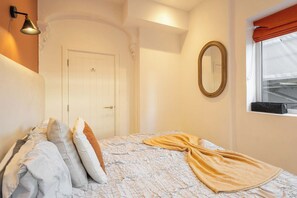 Un dormitori amb un llit, almoines, una làmpada de nit, un mirall i una finestra amb cortines.