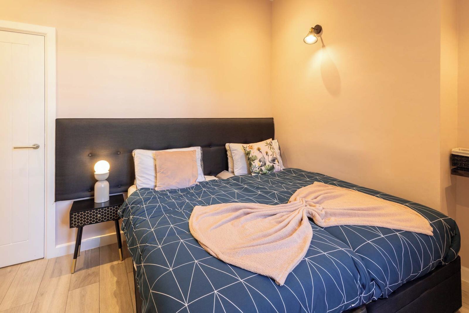 A neatly made bed with a geometric patterned bedspread, a bedside table with a lamp, and a wall-mounted light fixture.