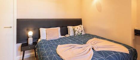 A neatly made bed with a geometric patterned bedspread, a bedside table with a lamp, and a wall-mounted light fixture.