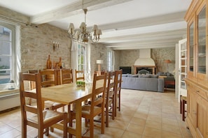 A dining area with a wooden table and chairs, a chandelier, and a stone wall. - Gîte l'Olivier - Domaine le Trabet (Moussoulens)