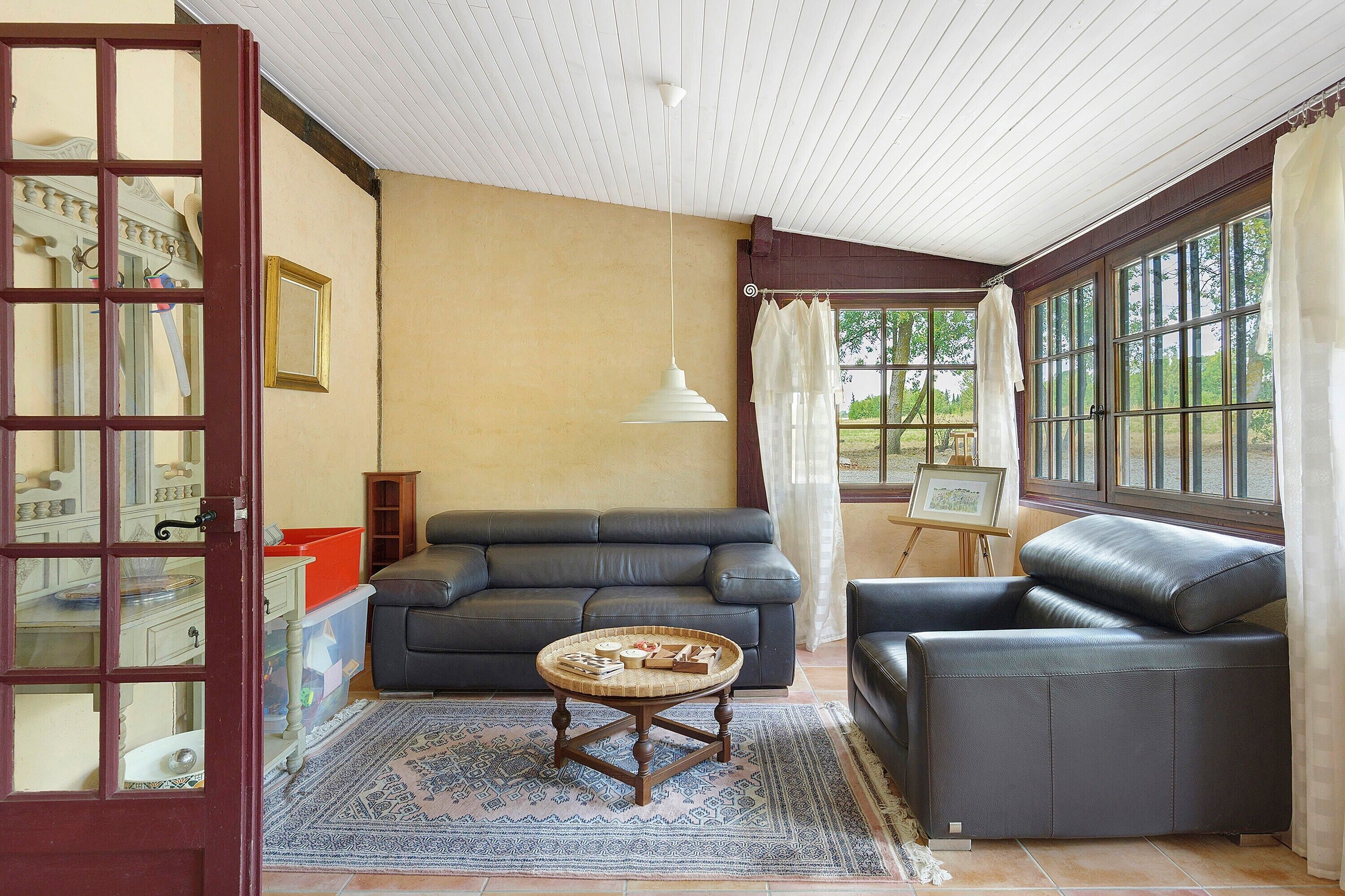 Un salon comprenant un canapé, un fauteuil et une petite table sur laquelle se trouve un panier. Il y a une fenêtre avec des rideaux, un tableau sur un chevalet et une porte avec un miroir.
