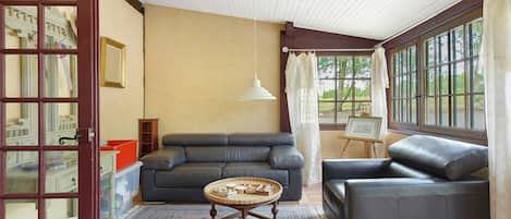Ein Wohnzimmer mit einer Couch, einem Sessel und einem kleinen Tisch mit einem Korb. Ein Fenster mit Vorhängen, ein Gemälde auf einem Lehrstuhl und eine Tür mit einem Spiegel sind ebenfalls vorhanden.