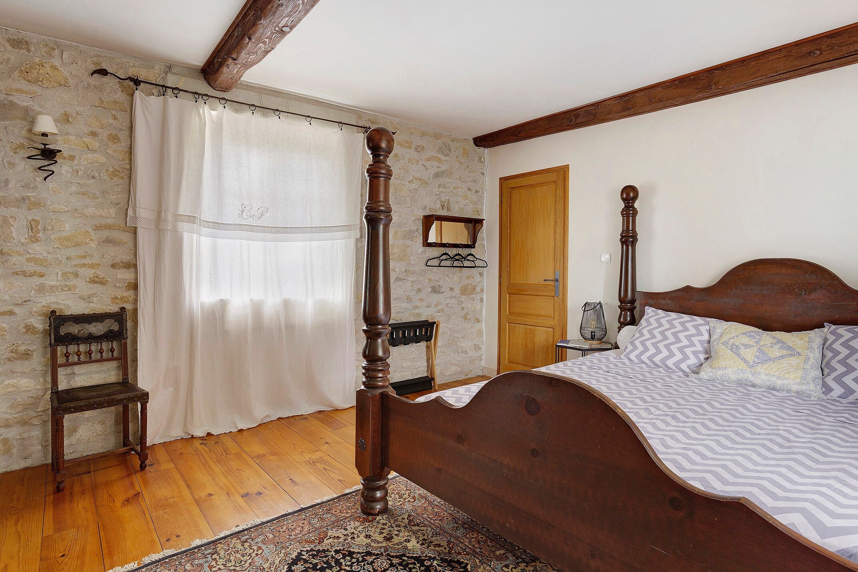 A bedroom with a four-poster bed, a wooden chair, a small table with a lamp, and a door.