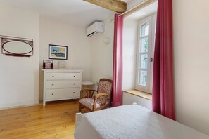 Ein Schlafzimmer mit Bett, Sessel, Kommode, einem Fenster mit Vorhängen und Wandaircondition.