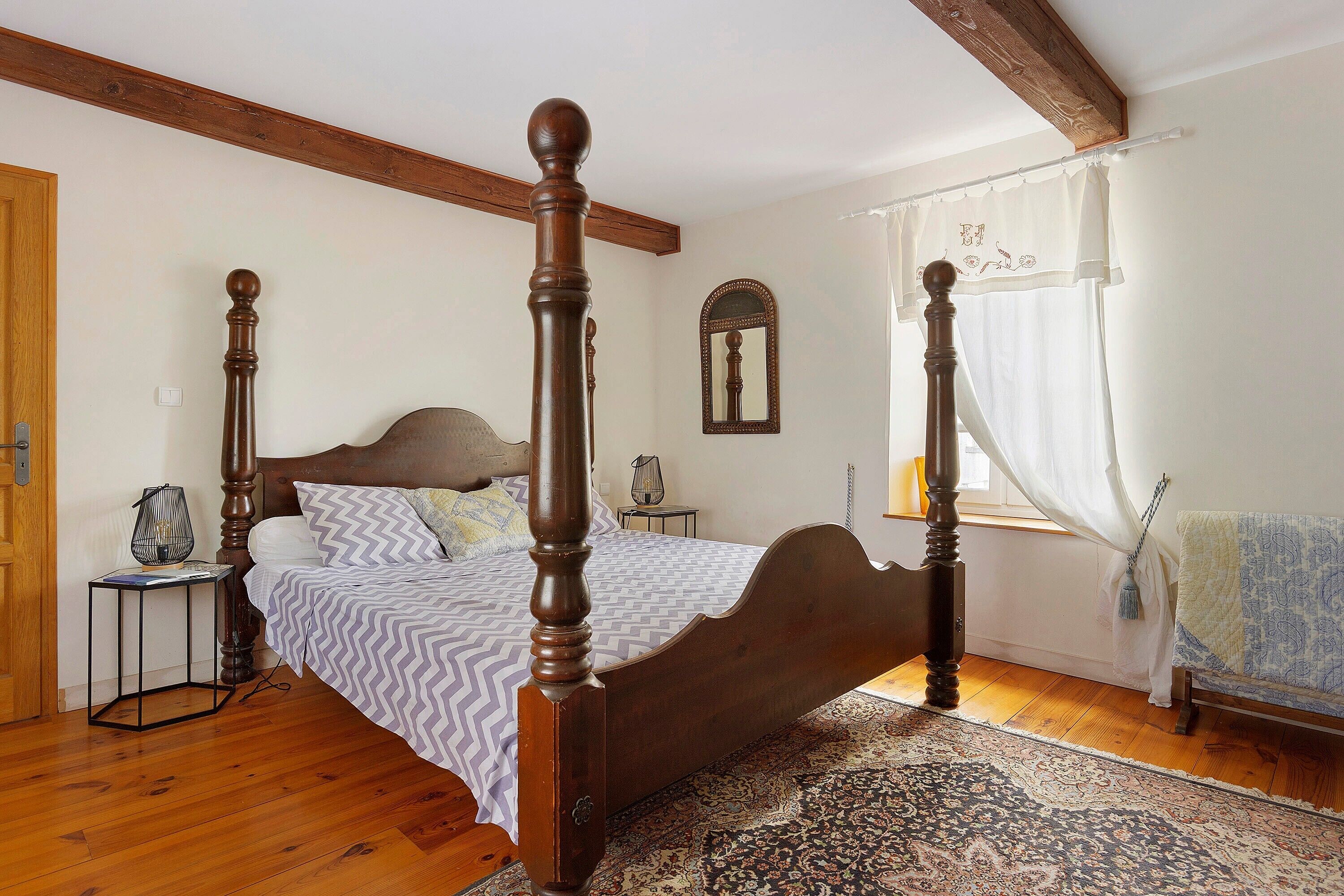 A bedroom with a four-poster bed, a nightstand, a mirror, and a window with curtains.
