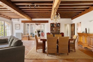Ein geräumiges Wohnzimmer mit hoher Decke, Holzbalken, einer Chandelüster und einem Essbereich mit Korbstühlen und einem Tisch.