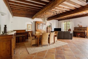 Ein geräumiges Wohnzimmer mit hoher Holzbalkendecke, einem Essbereich mit Korbstühlen, einem Klavier und einem Kamin.
