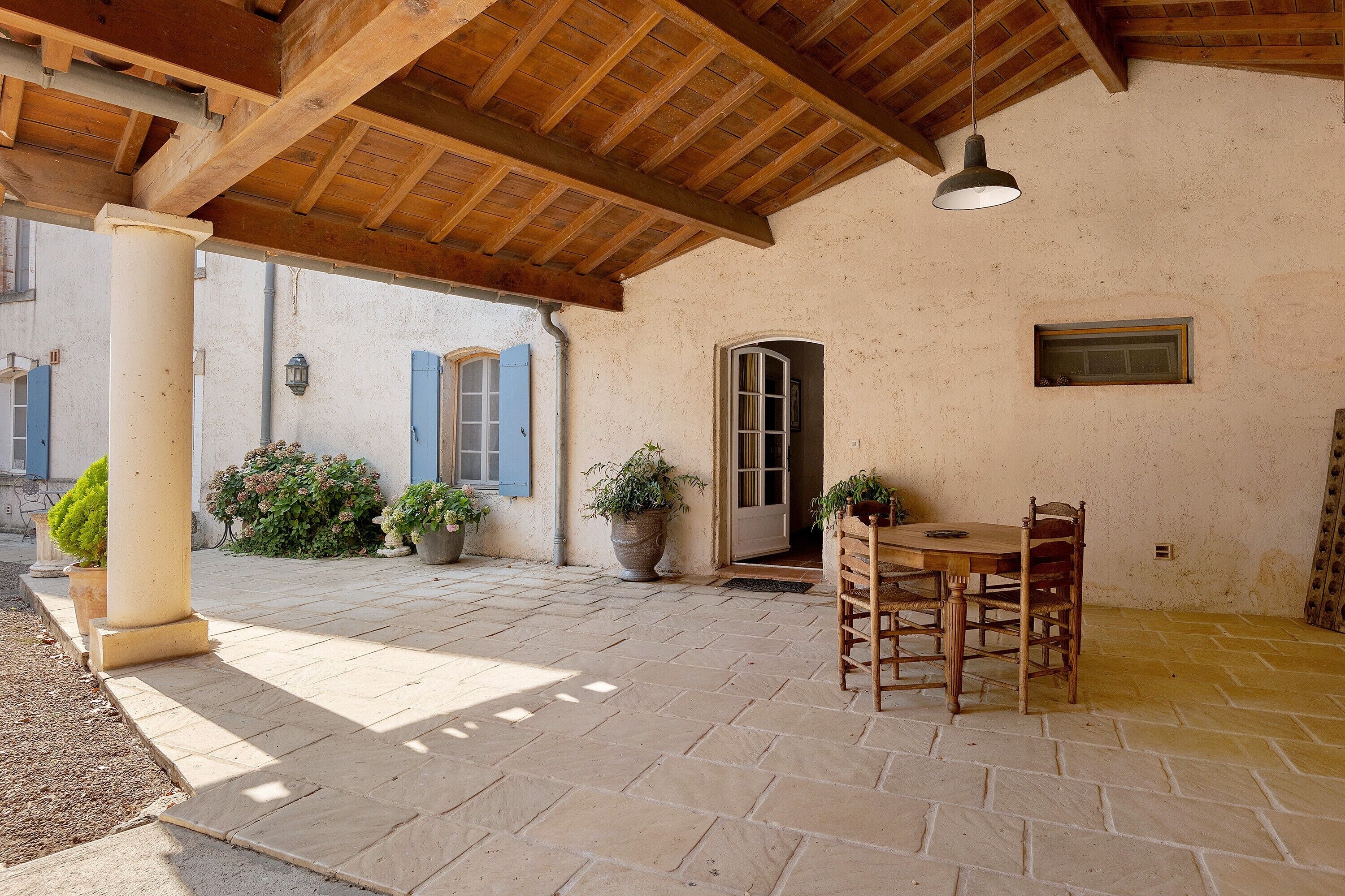 Une terrasse extérieure rustique, avec une table et des chaises en bois, des plantes en pot et une porte donnant sur un espace intérieur.