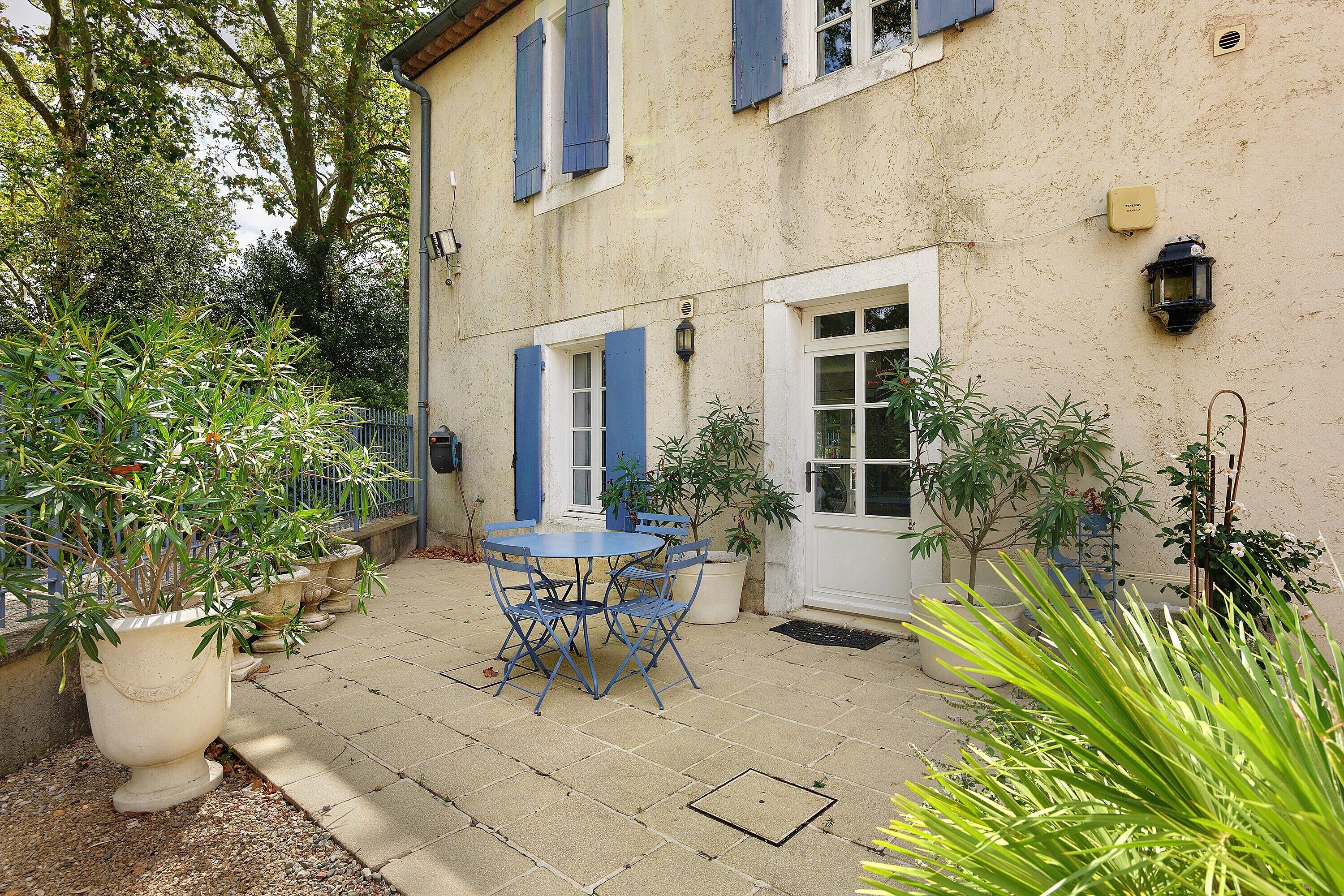 A courtyard with a table and chairs, surrounded by potted plants and a building with blue shutters.