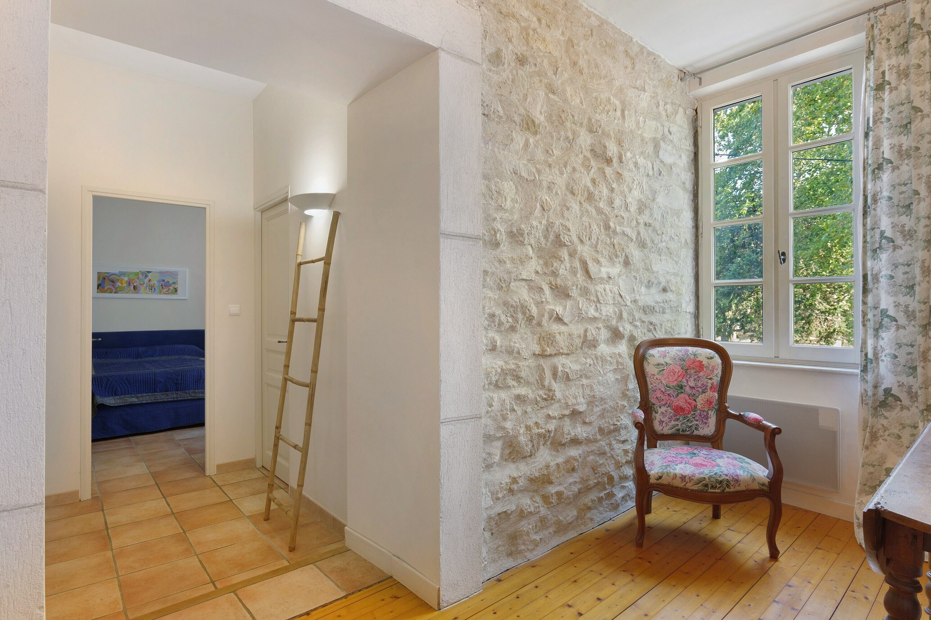 A room with a wooden floor, a chair with floral upholstery, a ladder, and a window with a view of greenery.