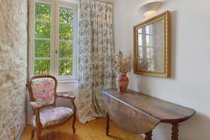 A room with a wooden chair, a table, a vase with dried plants, a mirror, and a window with curtains. - Domaine Le Trabet - Gîte Laurier (Moussoulens)