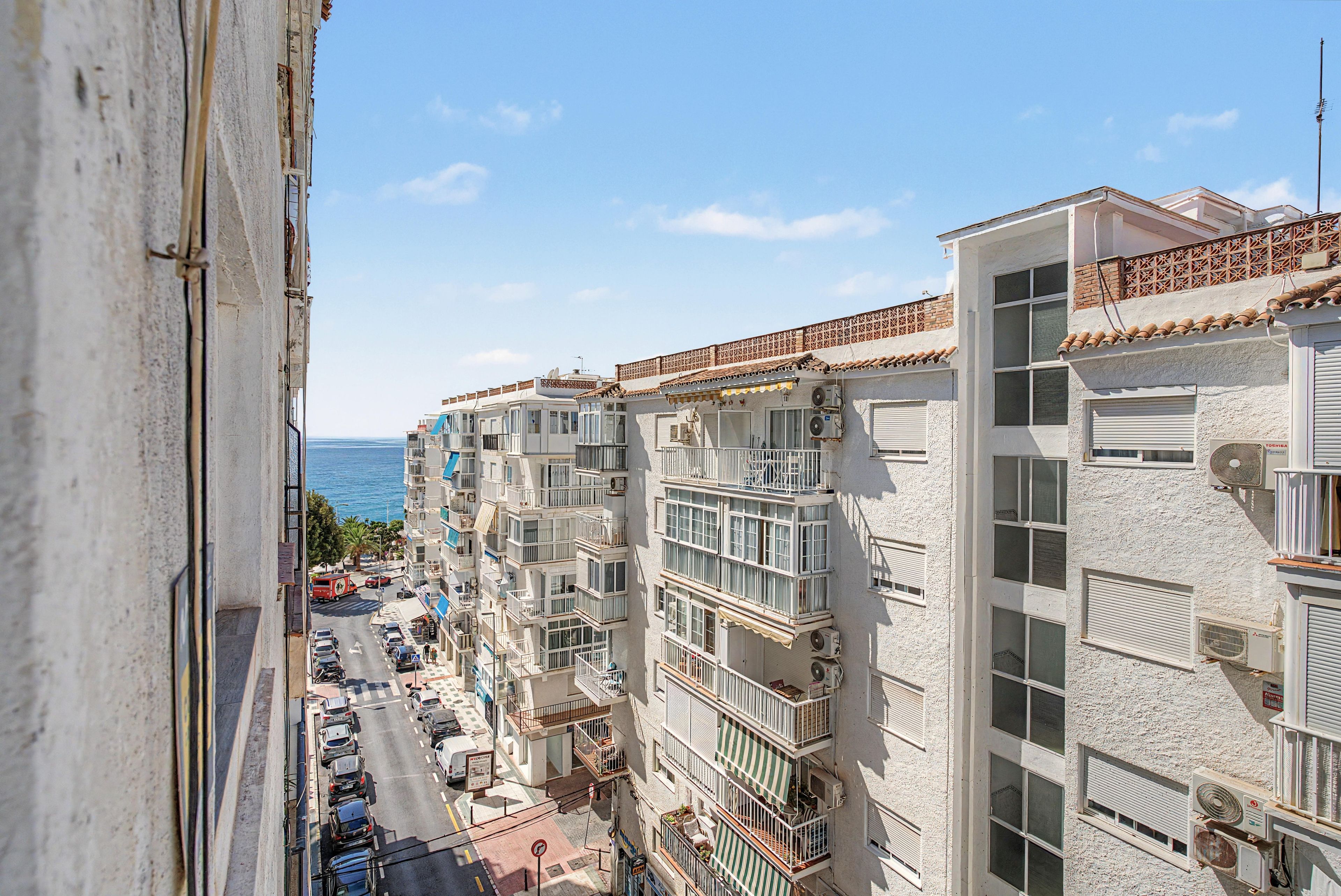 A multi-story residential building with balconies, a street view, and a clear view of the sea.