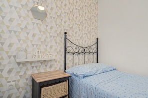 Ein Schlafzimmer mit Wand in geometrischem Muster, einem Bett mit blauen Musterbettwäsche, einem kleinen Nachttisch aus Holz und einem an der Wand befestigten Regal mit Dekorationen.