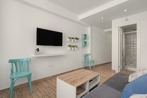 Ein modernes Wohnzimmer mit einem Flachbildfernseher, zwei smaragdgrünen Sesseln, einem Holztisch und einer kleinen, weißen Regal mit Pflanzen.