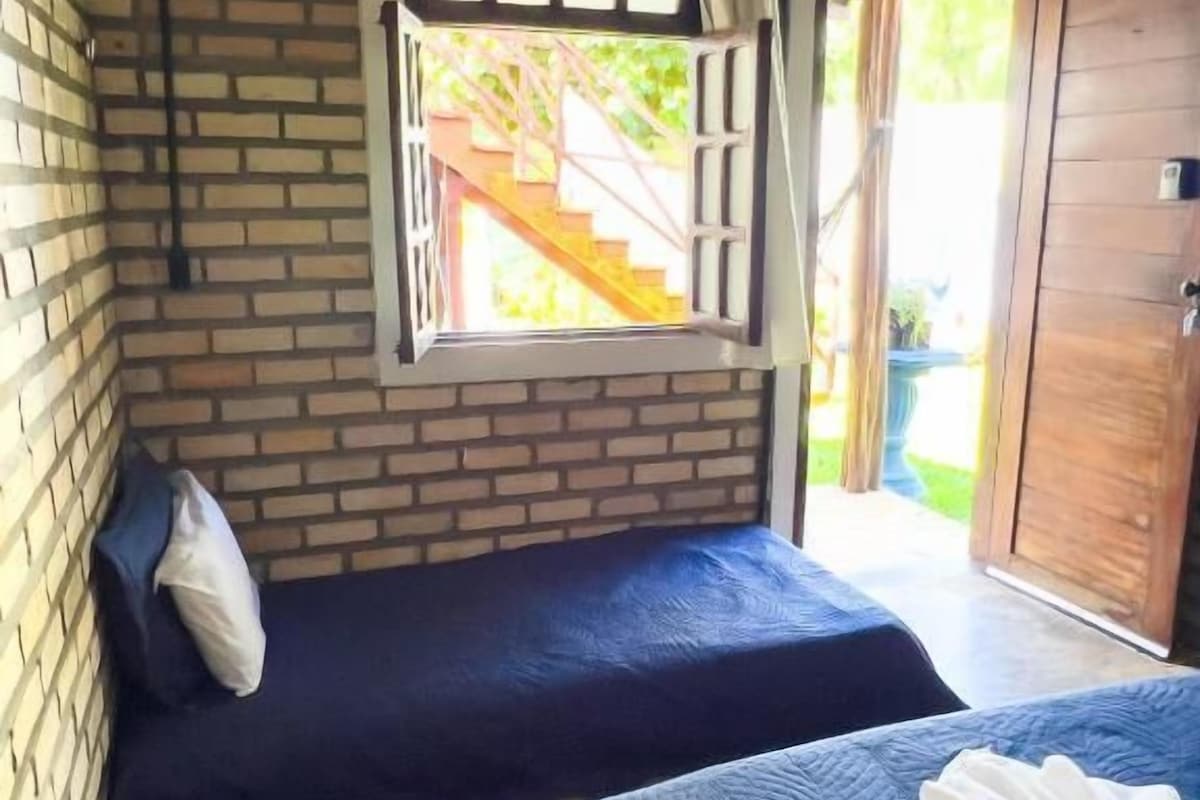A room with two beds, a window with curtains, a wooden door, and a brick wall.
