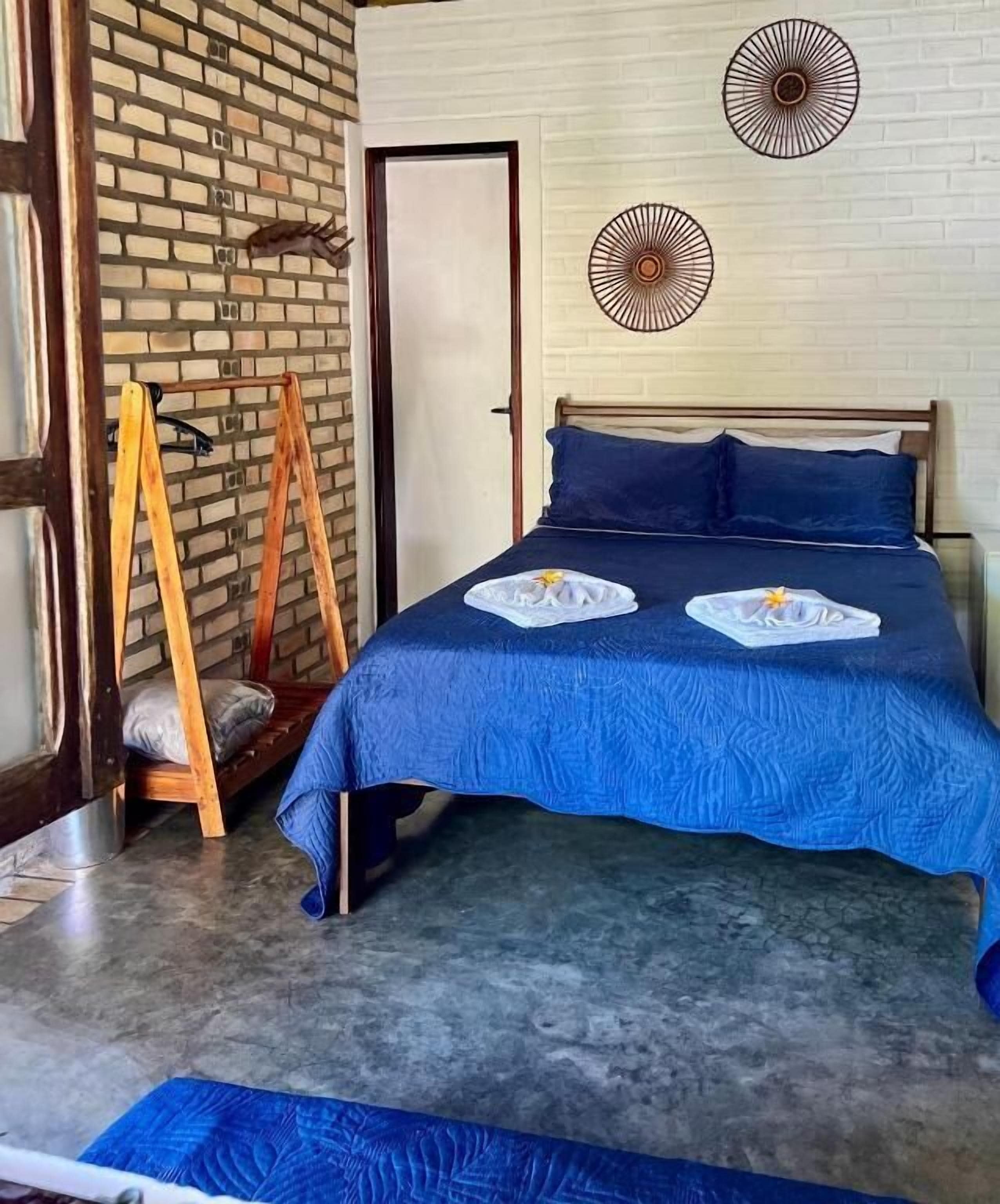 A bedroom with a brick wall, a bed with blue bedding, two wall-mounted fans, and a wooden ladder.