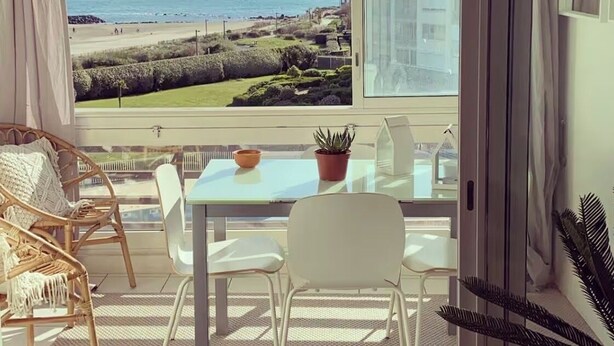 Une pièce dotée d’une grande fenêtre donnant sur une plage, d’un fauteuil en osier, d’une table blanche et d’une plante en pot.