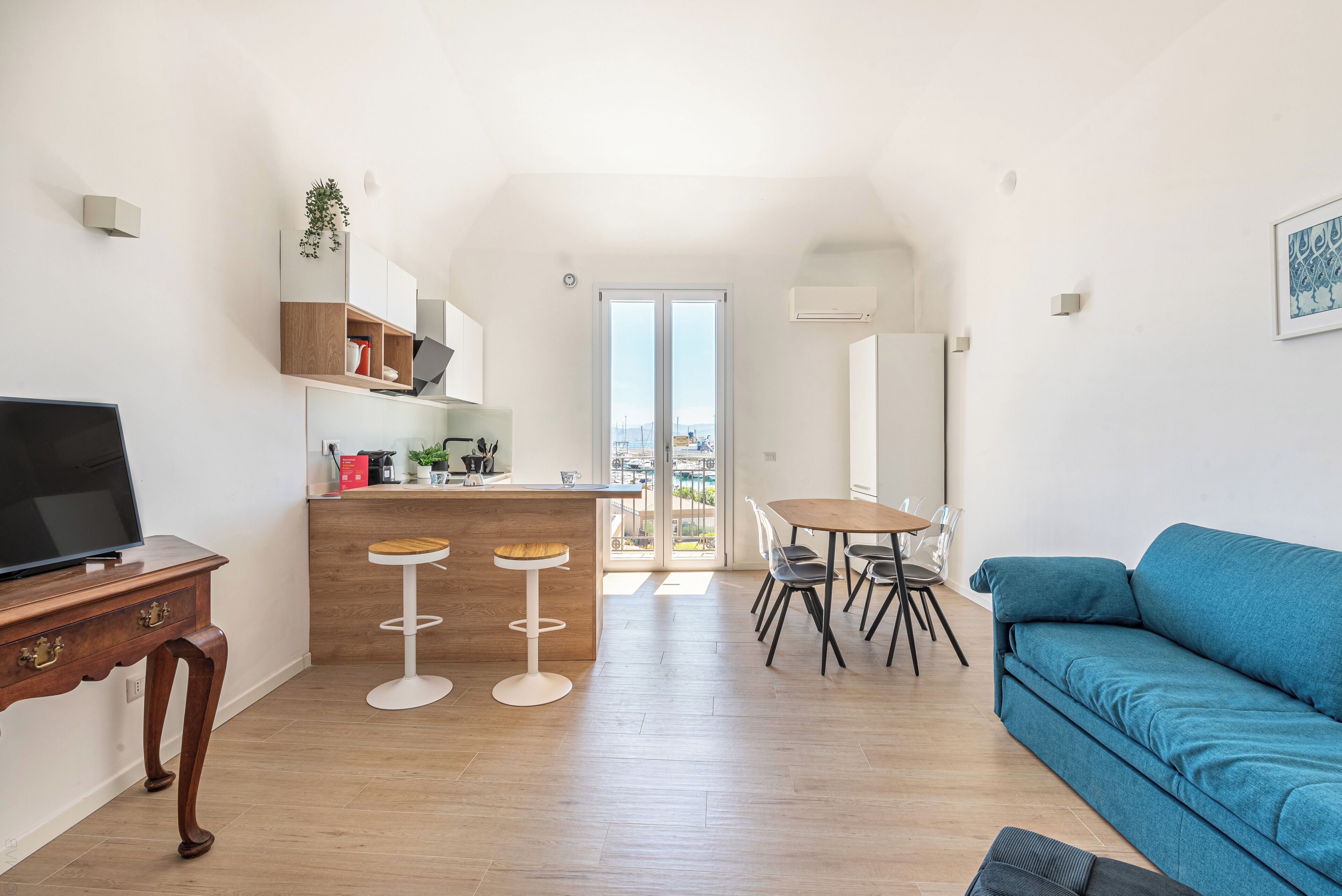 Un espacio habitable moderno con área de cocina, una mesa de comedor con sillas, un sofá azul y un televisor sobre un mueble de madera.