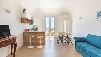 Un espacio habitable moderno con área de cocina, una mesa de comedor con sillas, un sofá azul y un televisor sobre un mueble de madera.