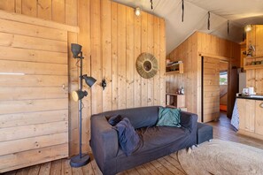 Interior acogedor de una cabaña con piso de madera, un sofá con cojines, una lámpara de pie y una cocina con armarios de madera.
